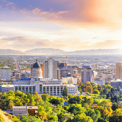 Aerial view of Salt Lake City, Utah during sunset. The foreground shows the Utah State Capitol building with its distinctive dome, surrounded by autumn trees with gold and green foliage. Downtown skyscrapers rise beyond the Capitol. In the background, the Wasatch mountain range stretches across the horizon under a colorful sky with pink and orange clouds.