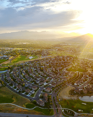 Aerial view of Cedar Hills, Utah at sunset or sunrise. The image shows a planned suburban neighborhood with curved streets and residential homes arranged in a circular pattern. A golf course with green fairways and small ponds is visible near the housing development. In the background, mountains rise against a golden-lit sky with scattered clouds, while the sun casts a warm glow across the valley. The perspective appears to be from a drone or elevated position overlooking the community with both developed residential areas and open spaces visible.